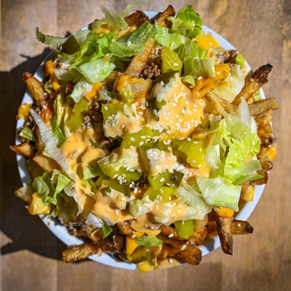 A close-up of loaded fries with cheese sauce, ground beef, lettuce, pickles, and sesame seeds.